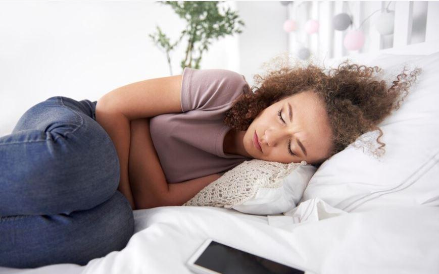 Žena leží na posteli, Drží se za břicho. Nepravidelná a bolestivá menstruace může značit neplodnost. Řada žen ale potíže podceňuje